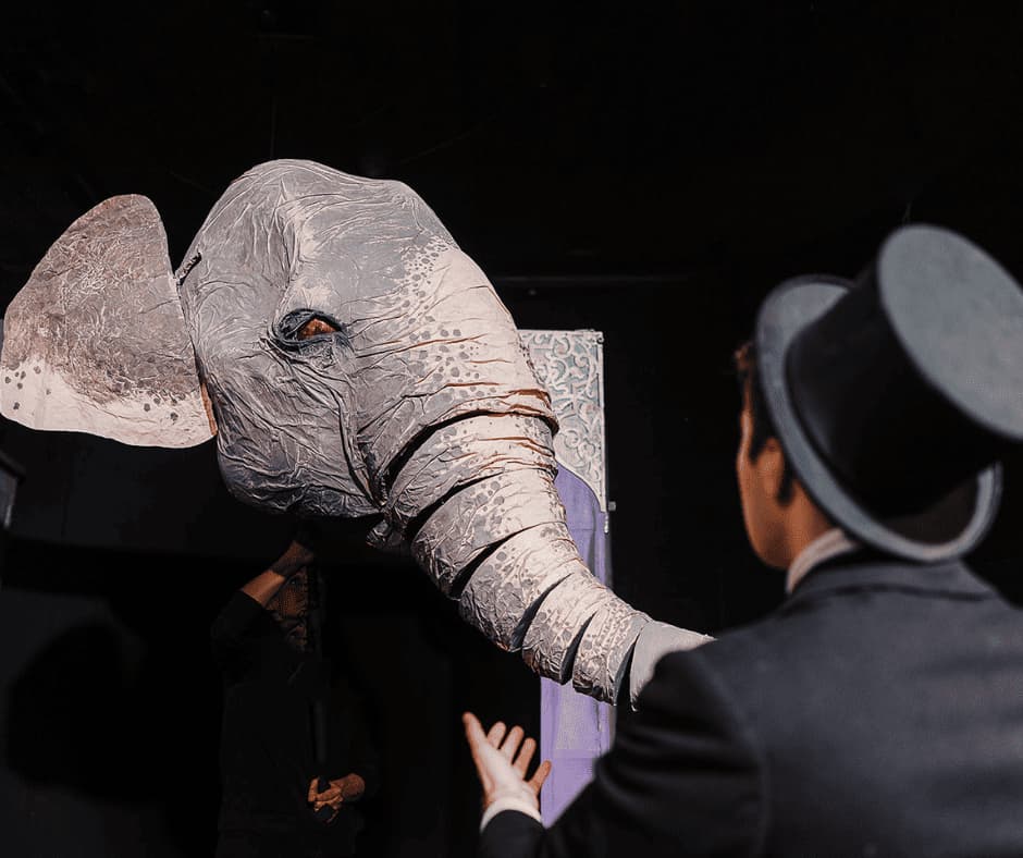 An elephant head puppet faces a man in a top hat