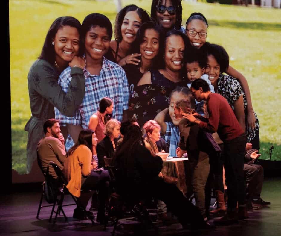 People seated and standing around a table interacting with each other with a large group photo behind them.