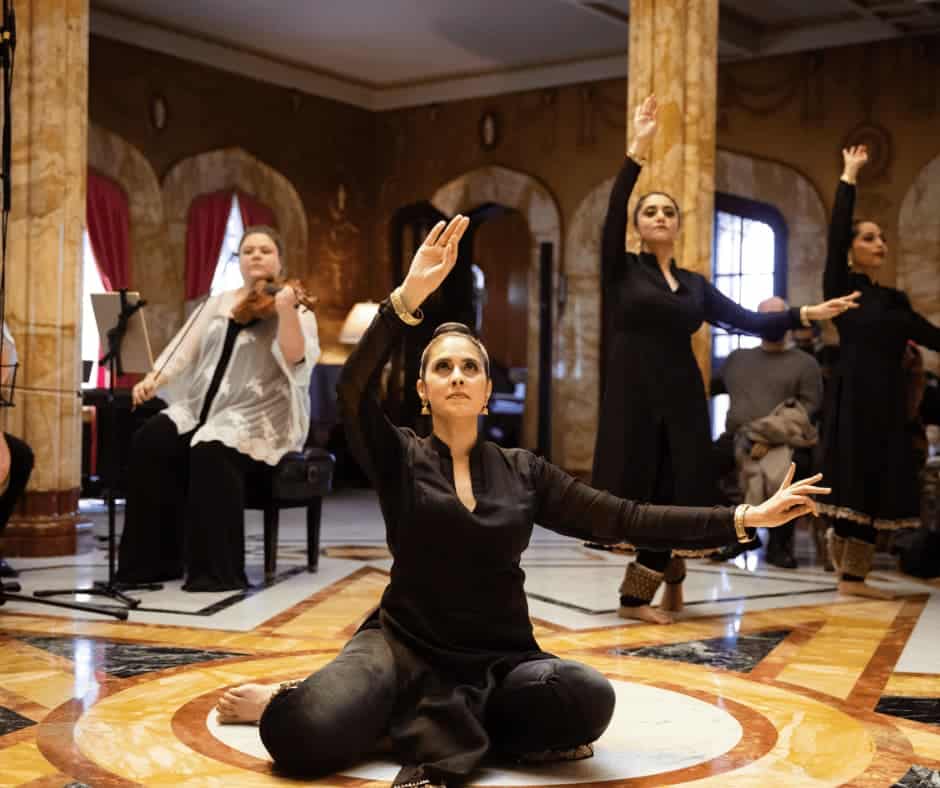 Three women dancing in a room with Middle Eastern decor. One is seated. A fourth woman plays a violin.