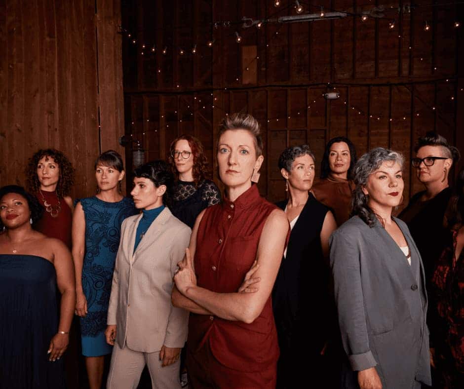 Ten women standing in a room with wooden walls and twinkle lights each is facing a different direction.