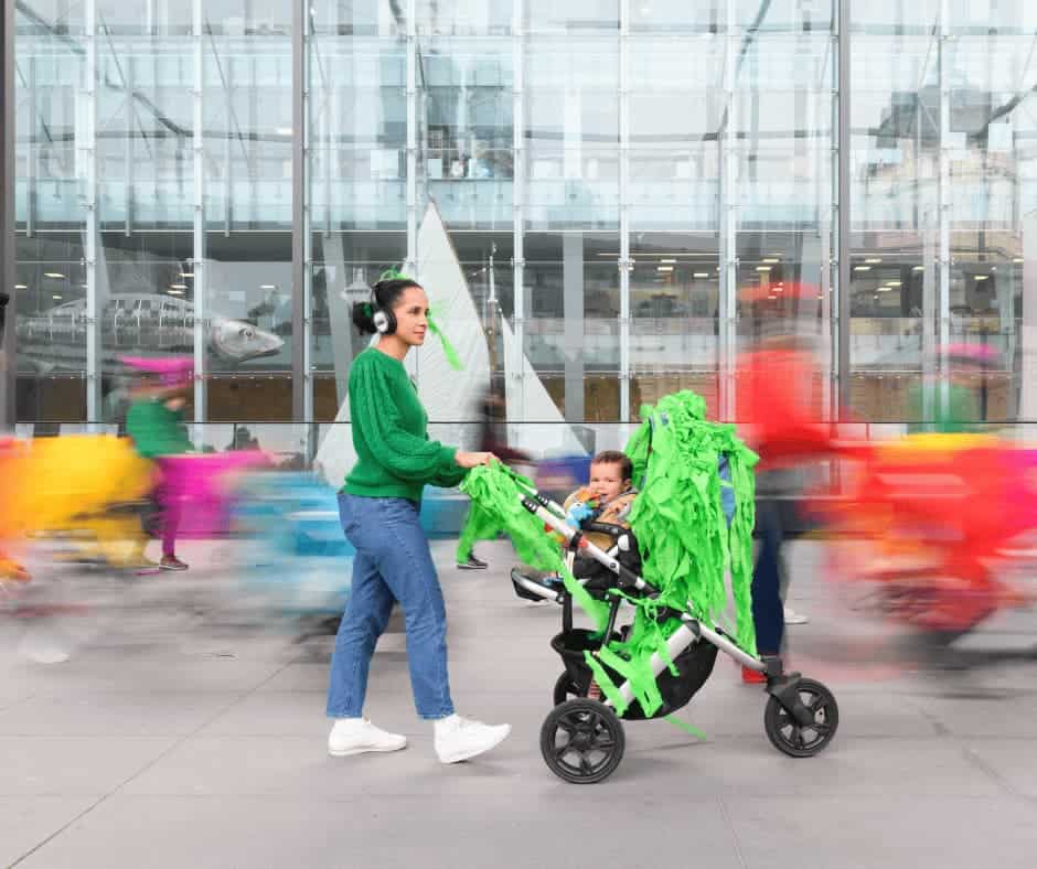 Woman pushing baby in a pram/stroller on a city sidewalk in front of glass building.