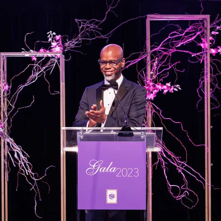 Black man wearing tuxedo standing behind a clear podium in front a flower wall