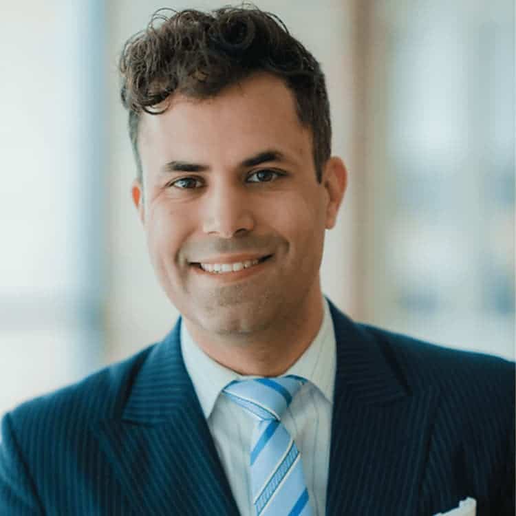 Man with dark short curly hair wearing a blue suit and tie