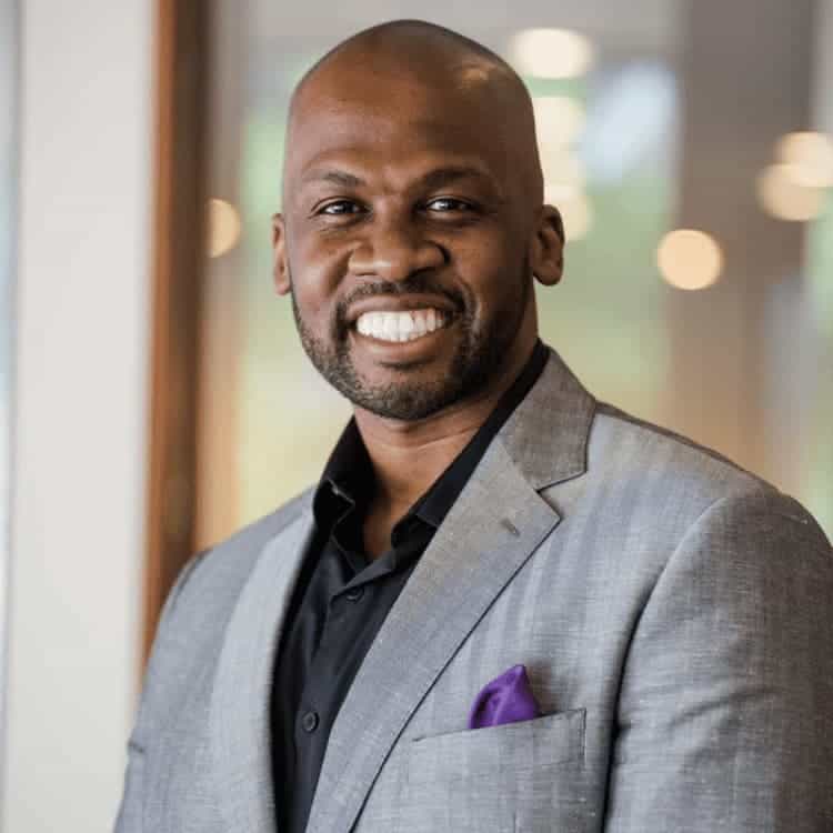 Black man smiling wearing a gray suit jacket and black shirt