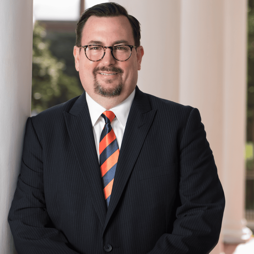 Chris is leaning against a white pillar wearing round dark glasses and a suit with a striped red and navy tie. He has light skin and short brown hair; photo by Auburn University Photographic Services.