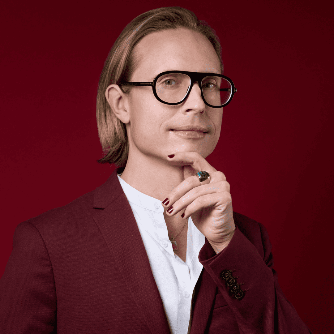 White femme person in aviator glasses, a burgundy suit and matching nails; photo by Simon Luethi.