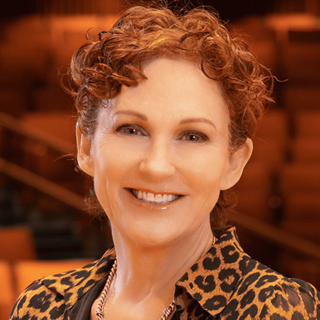Headshot of Beth, fair skin tone, short curly auburn hair, a leopard print dress, a gold necklace with theater seating in the background; photo by Peter Walker.