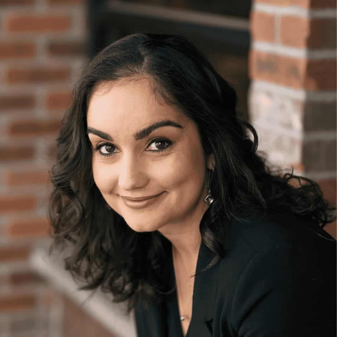 Aisha is smiling with medium length curly brown hair and olive skin. She is wearing a black shirt and is front of a blurred brick wall; photo by Stellar Propellar Studio.