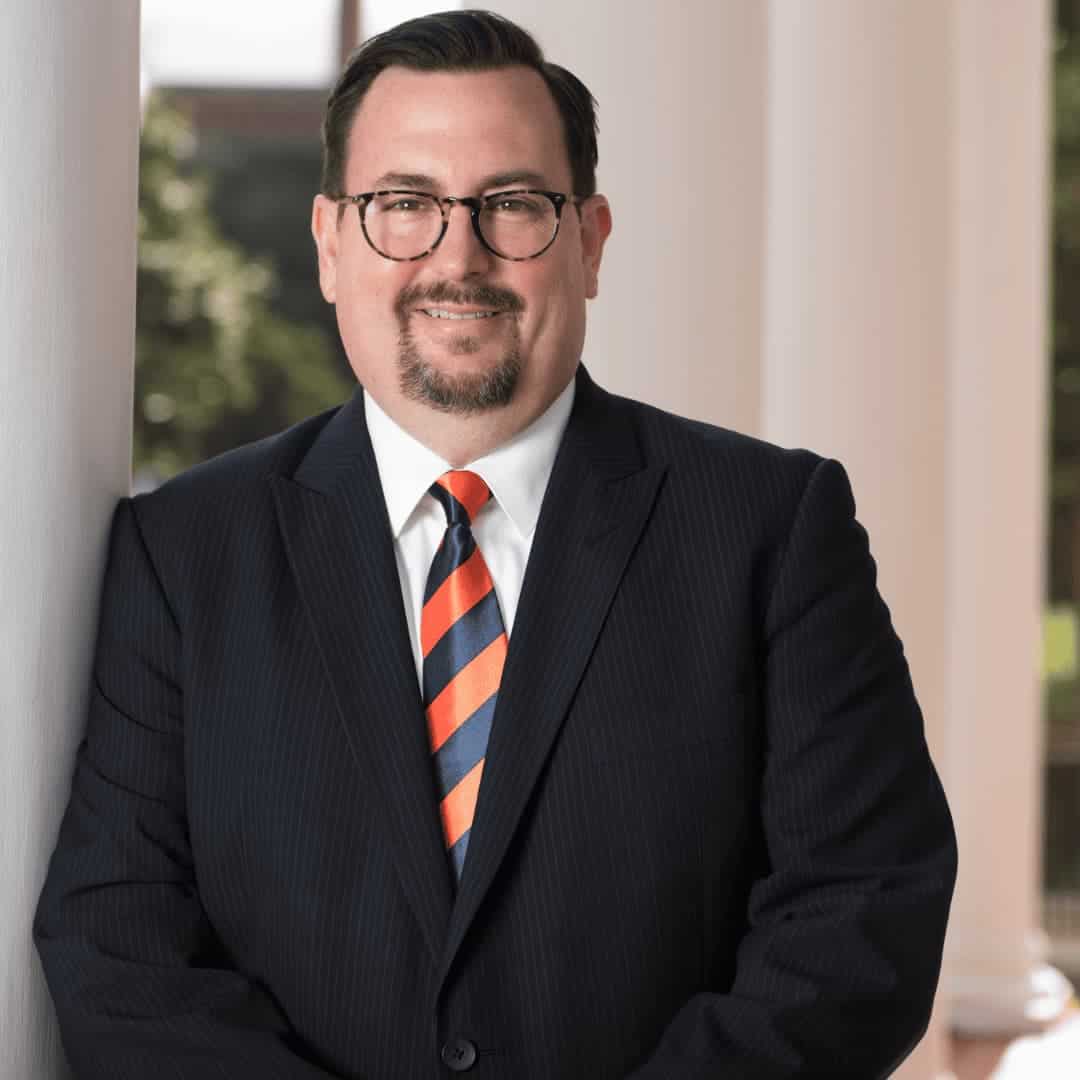 Chris is leaning against a white pillar wearing round dark glasses and a suit with a striped red and navy tie. He has light skin and short brown hair; photo by Auburn University Photographic Services.