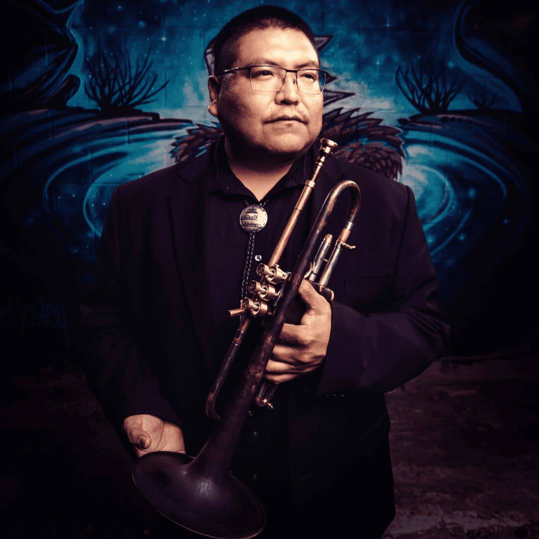 Headshot of Delbert, brown skin tone, with short black hair and glasses, wearing a black suite over a black buttoned shirt, holding a dark brown trumpet with dark blue graffiti art behind him as a background; photo by Maurice Johnson.