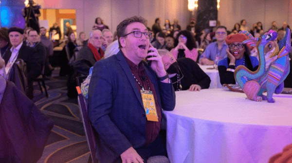 APAP member during an APAP|NYC 2024 session making a surprised face focusing on the main stage at their table. Background is filled with other APAP|NYC 2024 conference attendees who are also engrossed with what's going onstage.